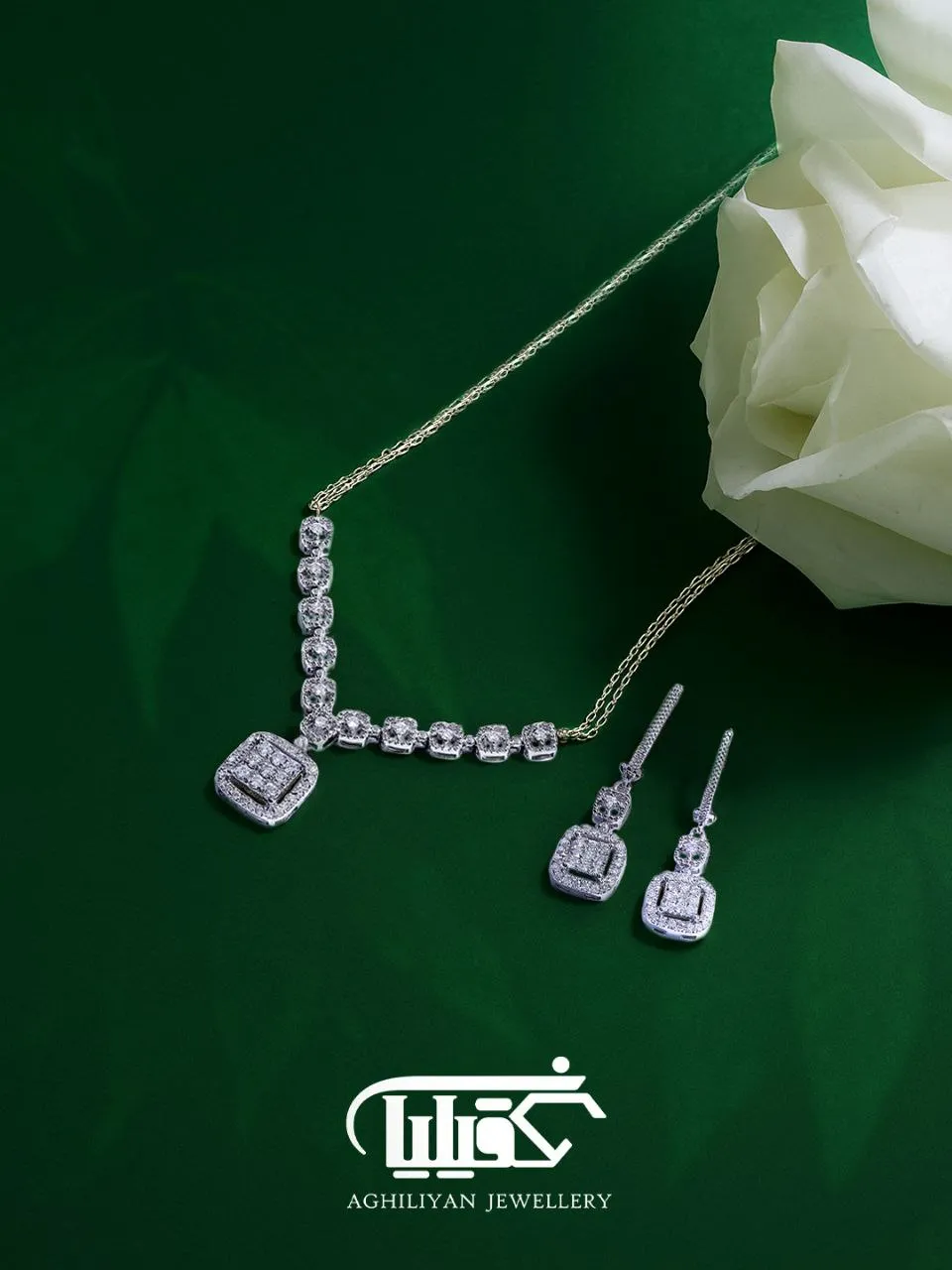 diamond-jewelry-set-3768-aghiliyan نیم ست جواهر الماس تراش برلیان کد 3768 شامل گردنبند و گوشواره آویز طلای سفید روی پسزمینه سبز در کنار گل رز سفید